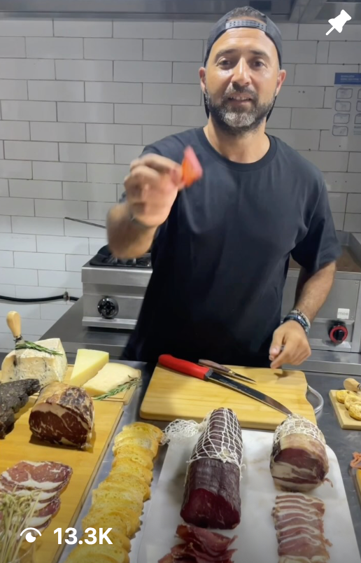 Man in a kitchen holding a piece of meat above a counter with various meats and cheeses.