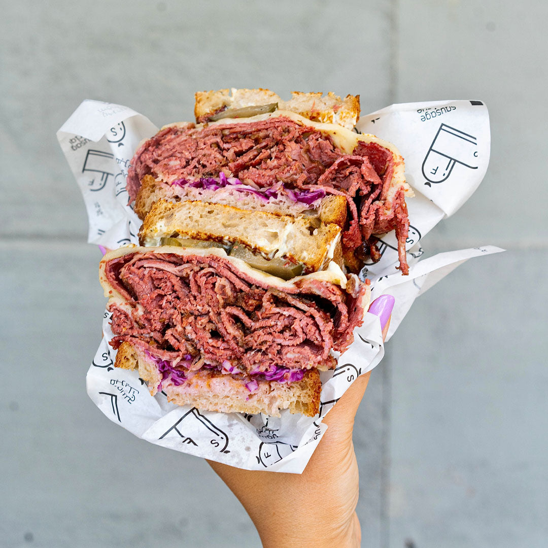 Half-eaten sandwich with pastrami beef and pickles on a piece of paper with graphics, held against a gray background.