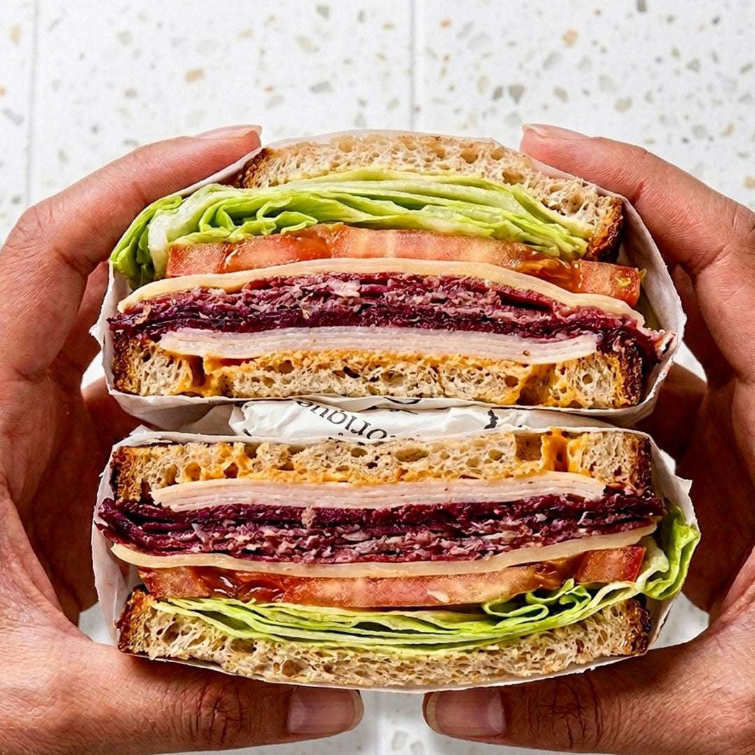 Hands holding a halved sandwich with visible layers of bread, meat, and vegetables.