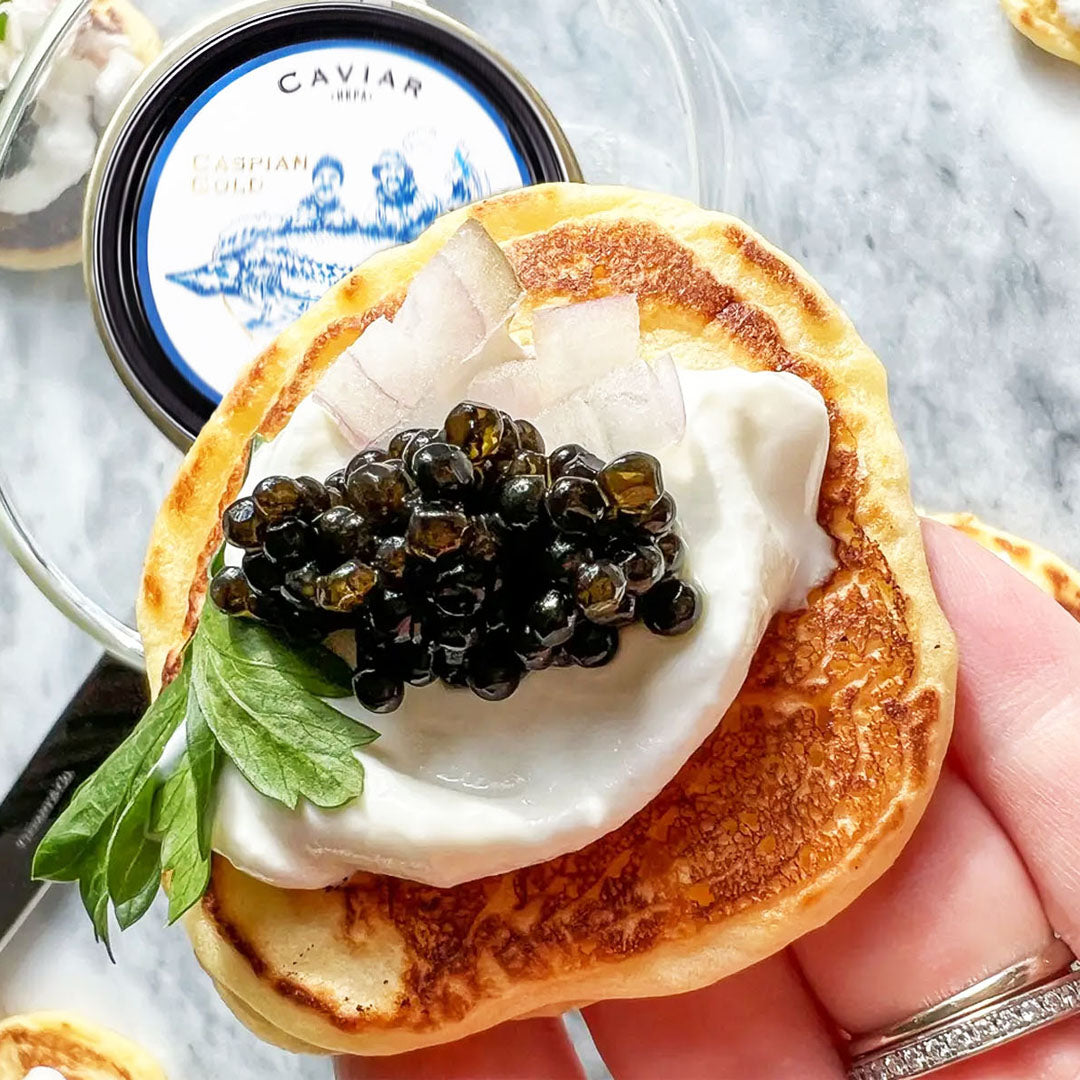 Small pancake with caviar and sour cream held by a hand, with a container of caviar in the background.