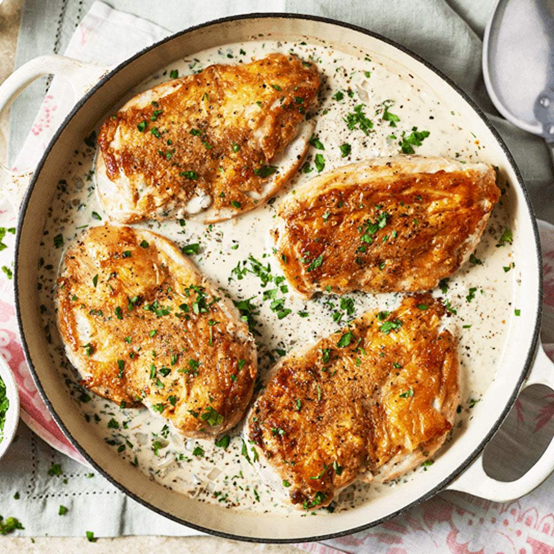 Pan of chicken with a creamy sauce and herbs on a patterned tablecloth.