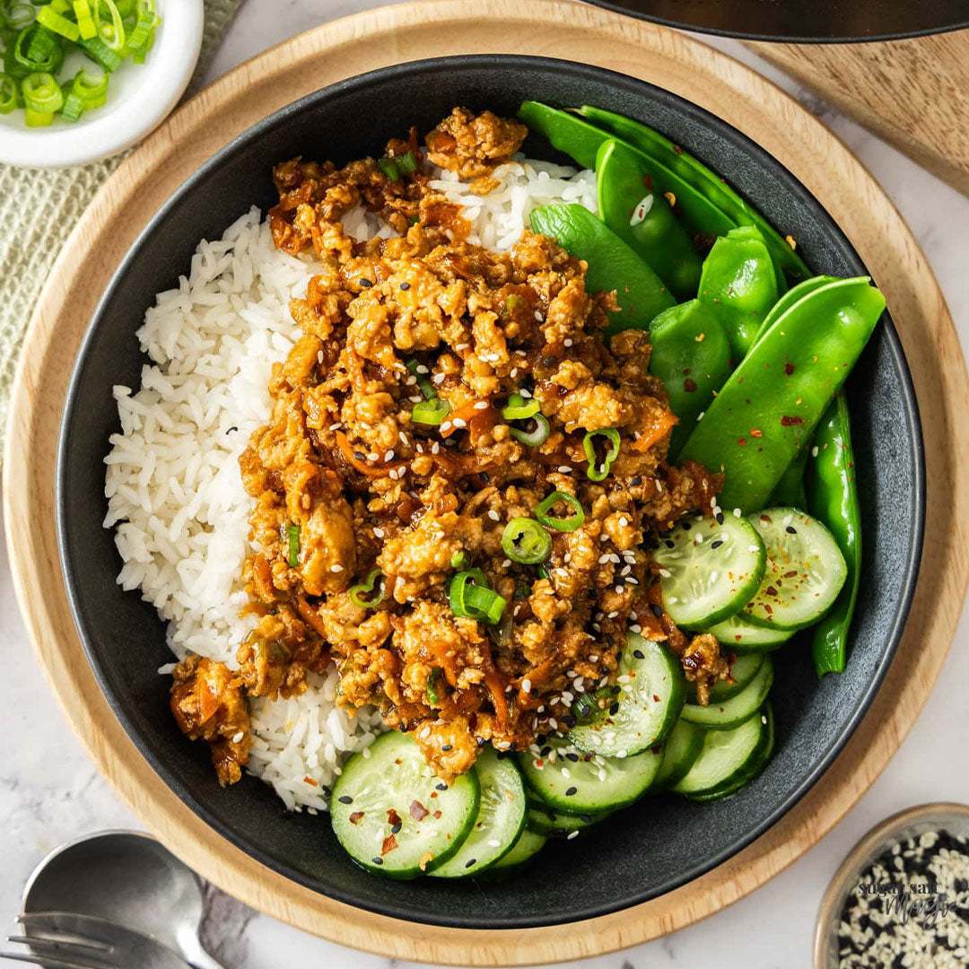 Bowl of rice with ground meat, snap peas, and cucumbers on a wooden table.