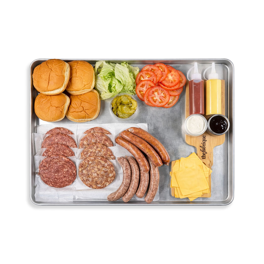 Metal tray with hamburger buns, sausages, cheese, tomatoes, and condiments on a white background.