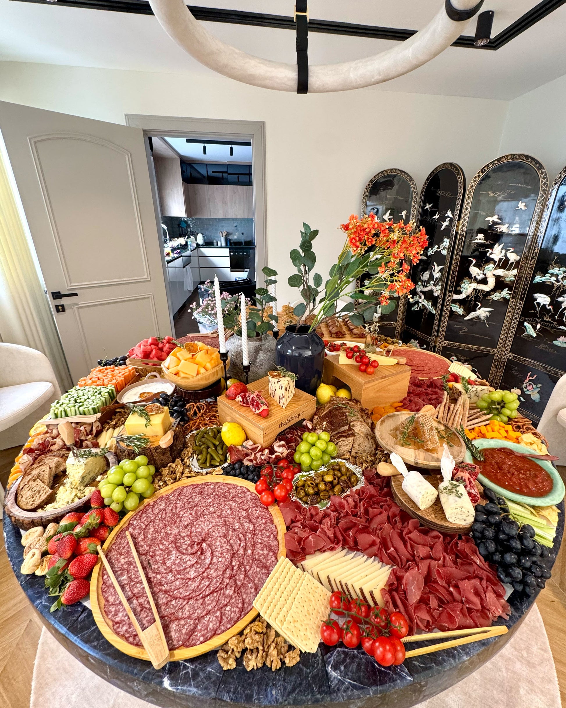 Round charcuterie board with various meats, cheeses, fruits, and vegetables on a dining table.