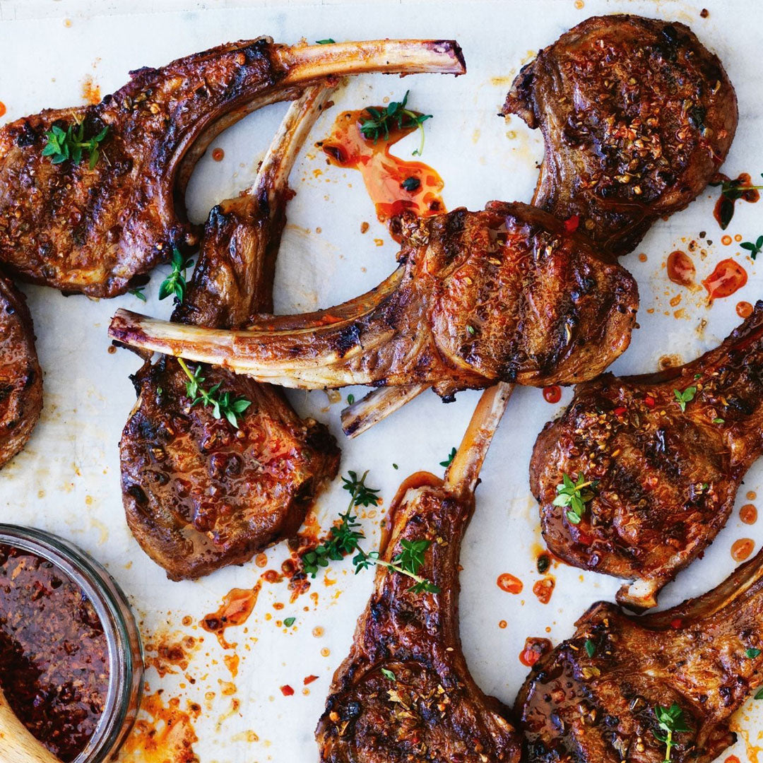 Grilled lamb chops on a white surface with a small bowl of sauce.