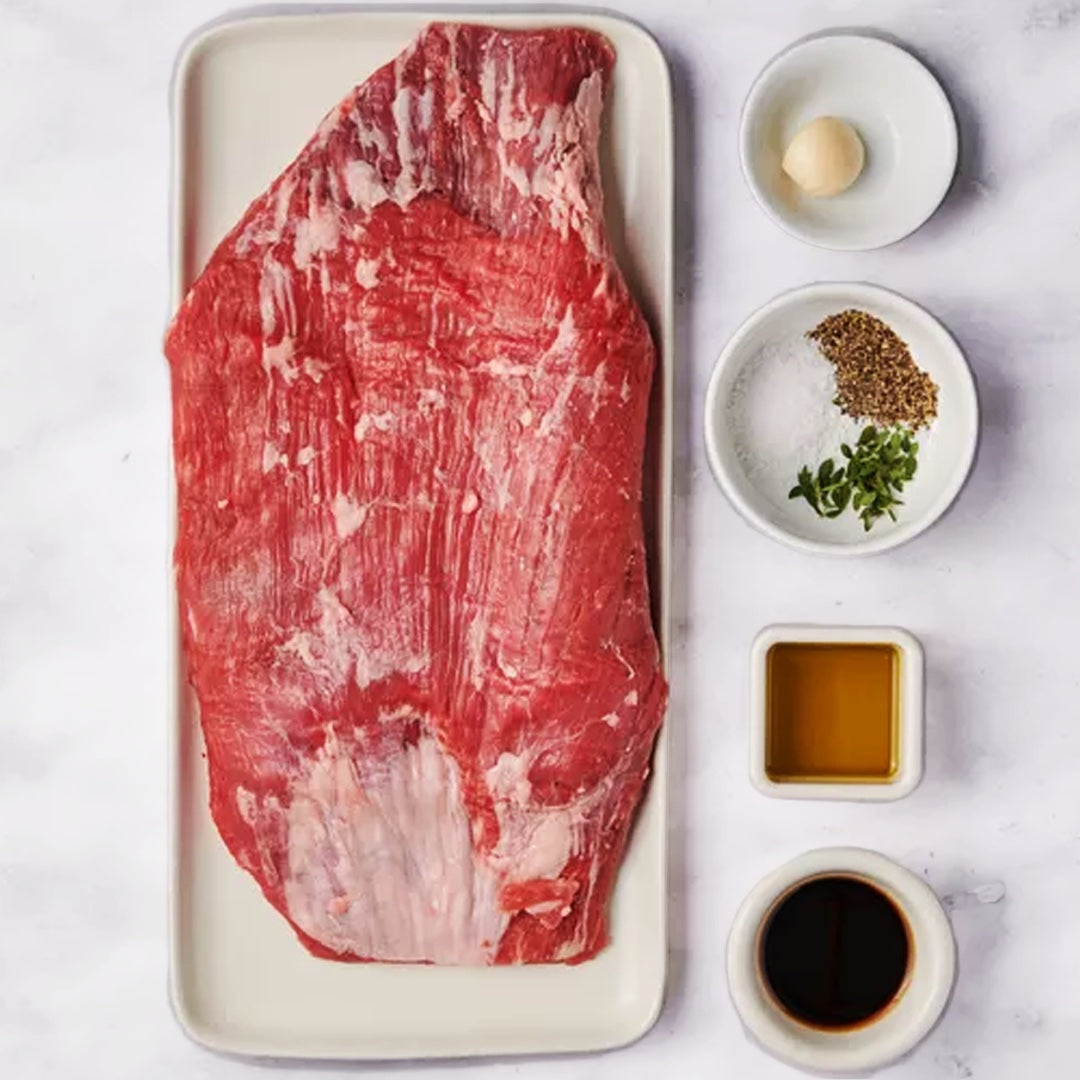 Raw flank steak on a white plate with seasonings and sauces on a light background