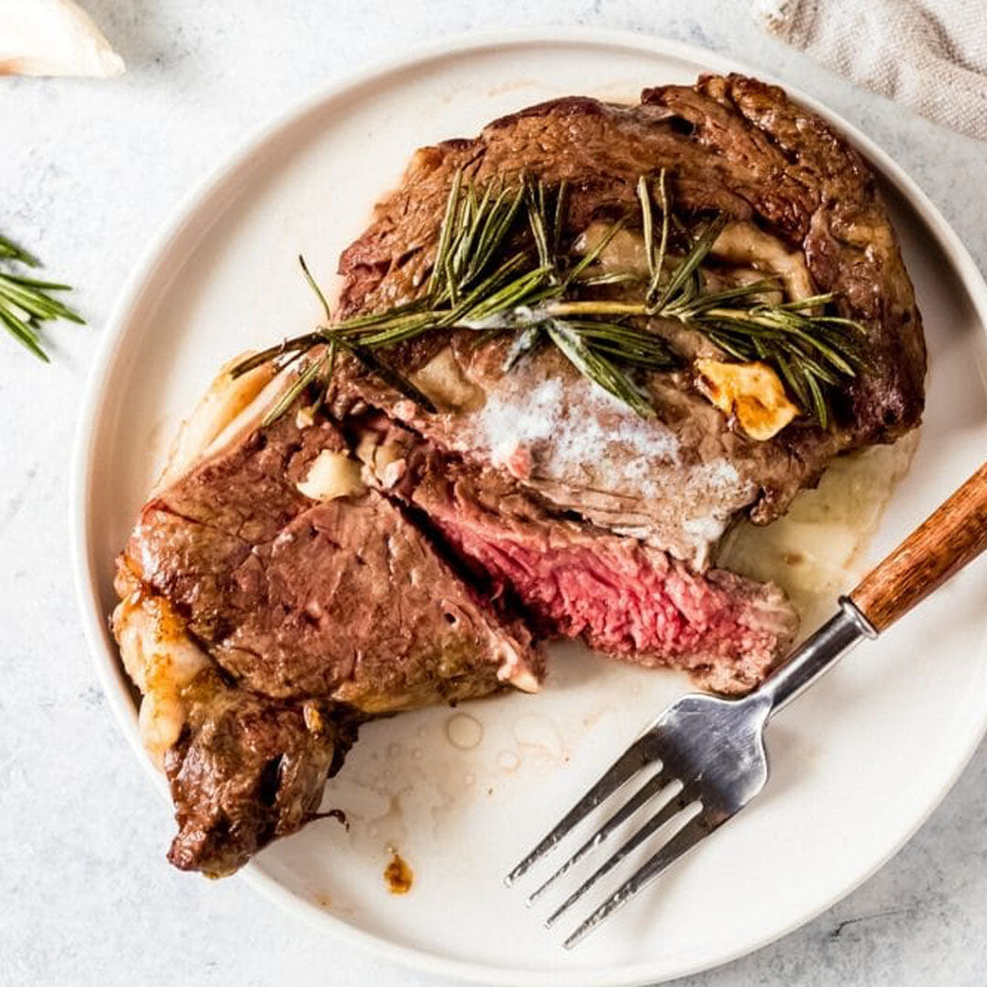 Steak with rosemary on a white plate with a fork, on a light gray surface.