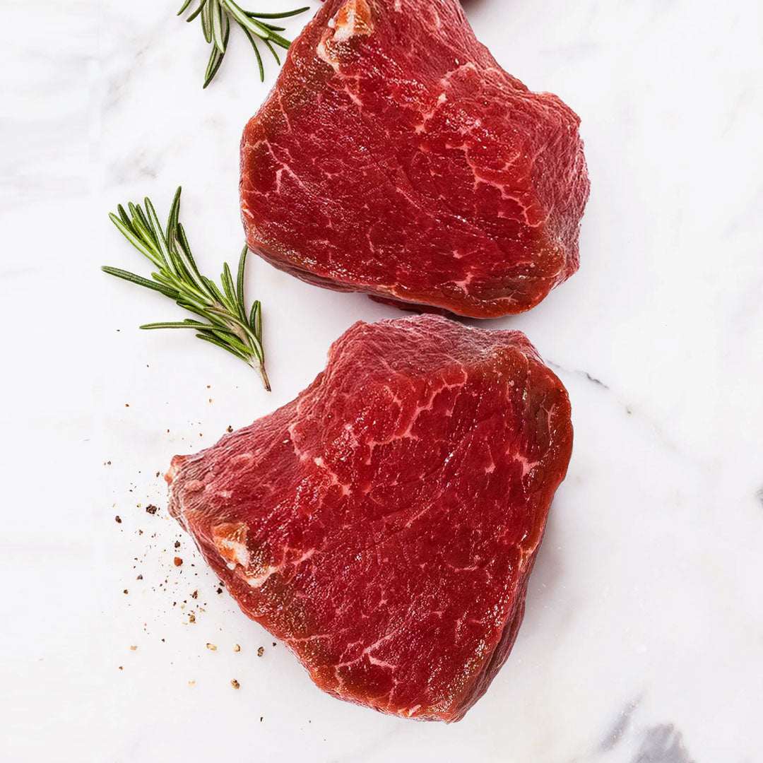 Two pieces of raw red meat with rosemary on a white surface