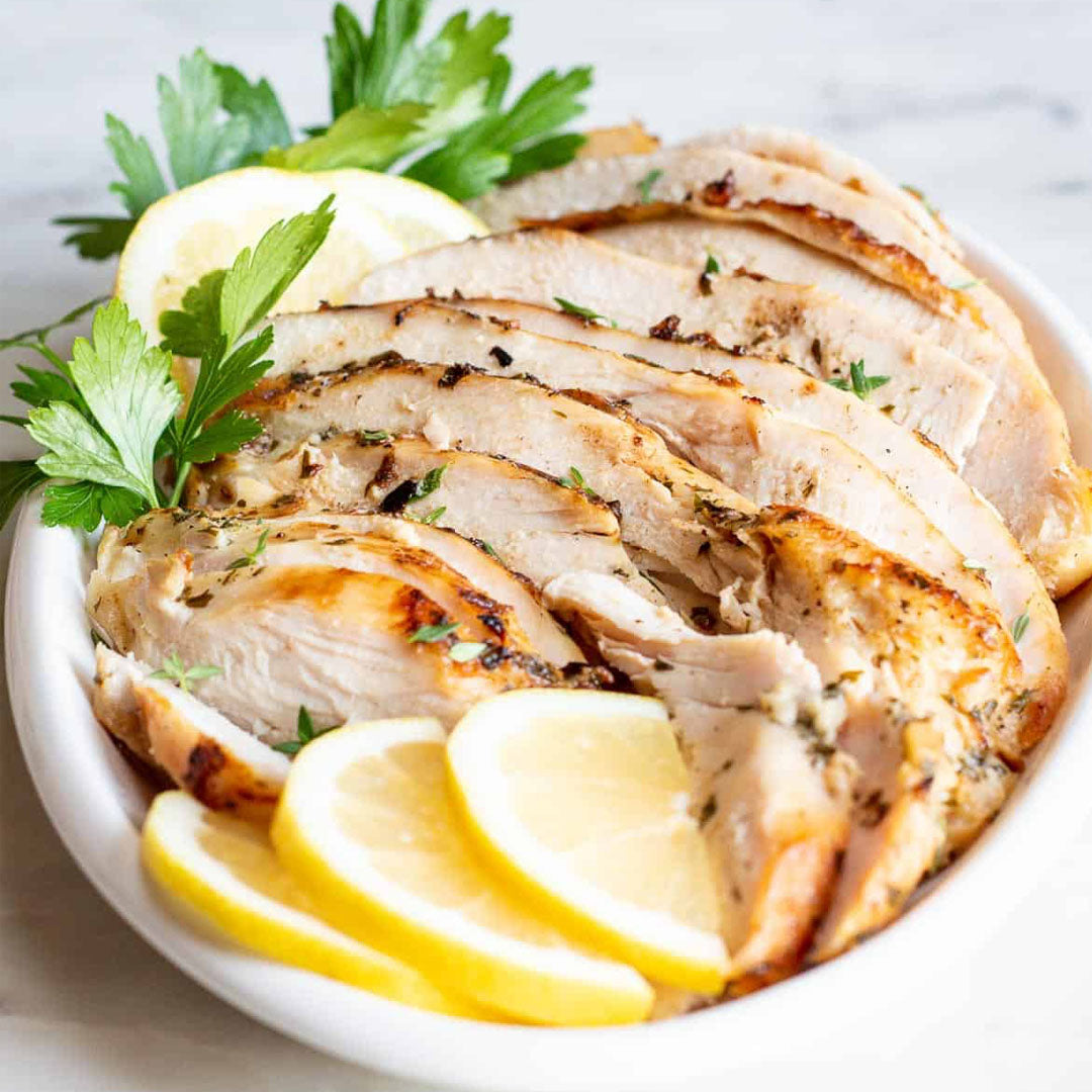 Sliced roasted chicken breast with lemon wedges and parsley on a white plate.
