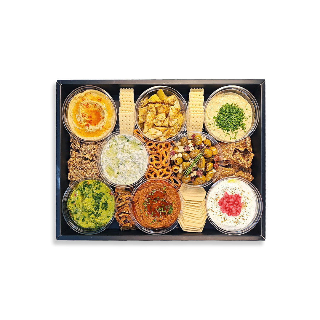A gourmet snack and appetizer box presented in a black rectangular tray on a white background. The box contains eight small clear bowls of dips and spreads, including hummus, tzatziki, baba ghanoush, tabbouleh, and salad, alongside crackers, pretzels, and olives.