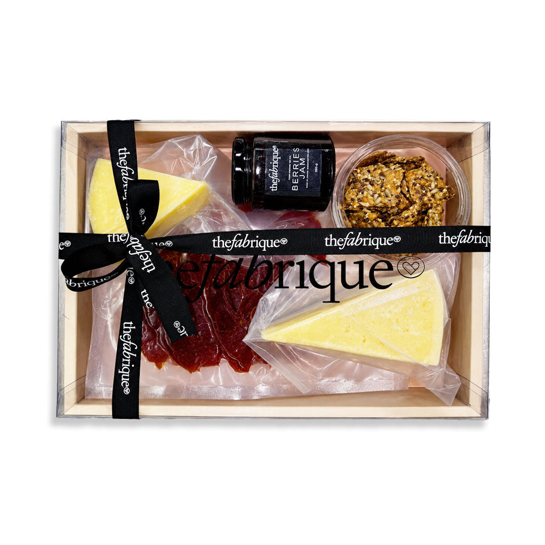 A gourmet cheese and charcuterie gift box from 'thefabrique' in a light wood tray, featuring two wedges of cheese, a dark red jam, a jar of preserves, a small bowl of granola, and a black ribbon printed with the brand name.