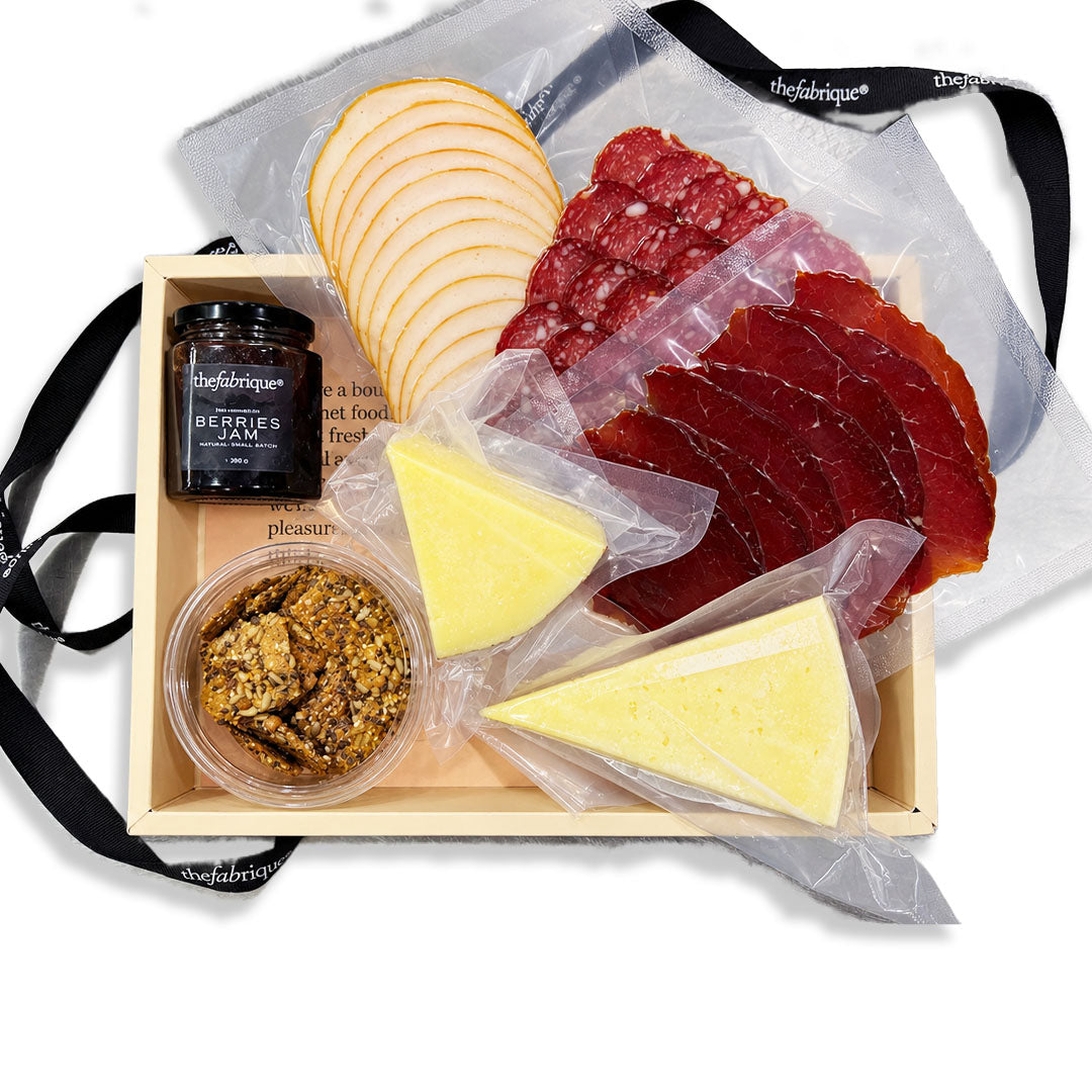 A charcuterie board gift set in a light wood tray, featuring sliced cured meats, two wedges of cheese, a small jar of honey, and a bowl of nuts, arranged with a clear plastic bag and black ribbon on a white background.