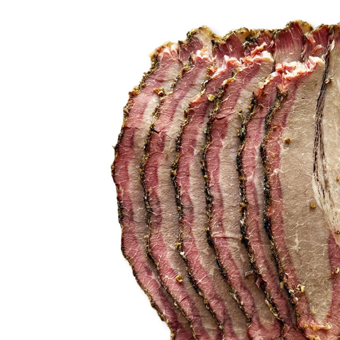 Close-up of sliced cured meat with pink-red interior and pale fat layers, topped with dark seasoning, on a white background.