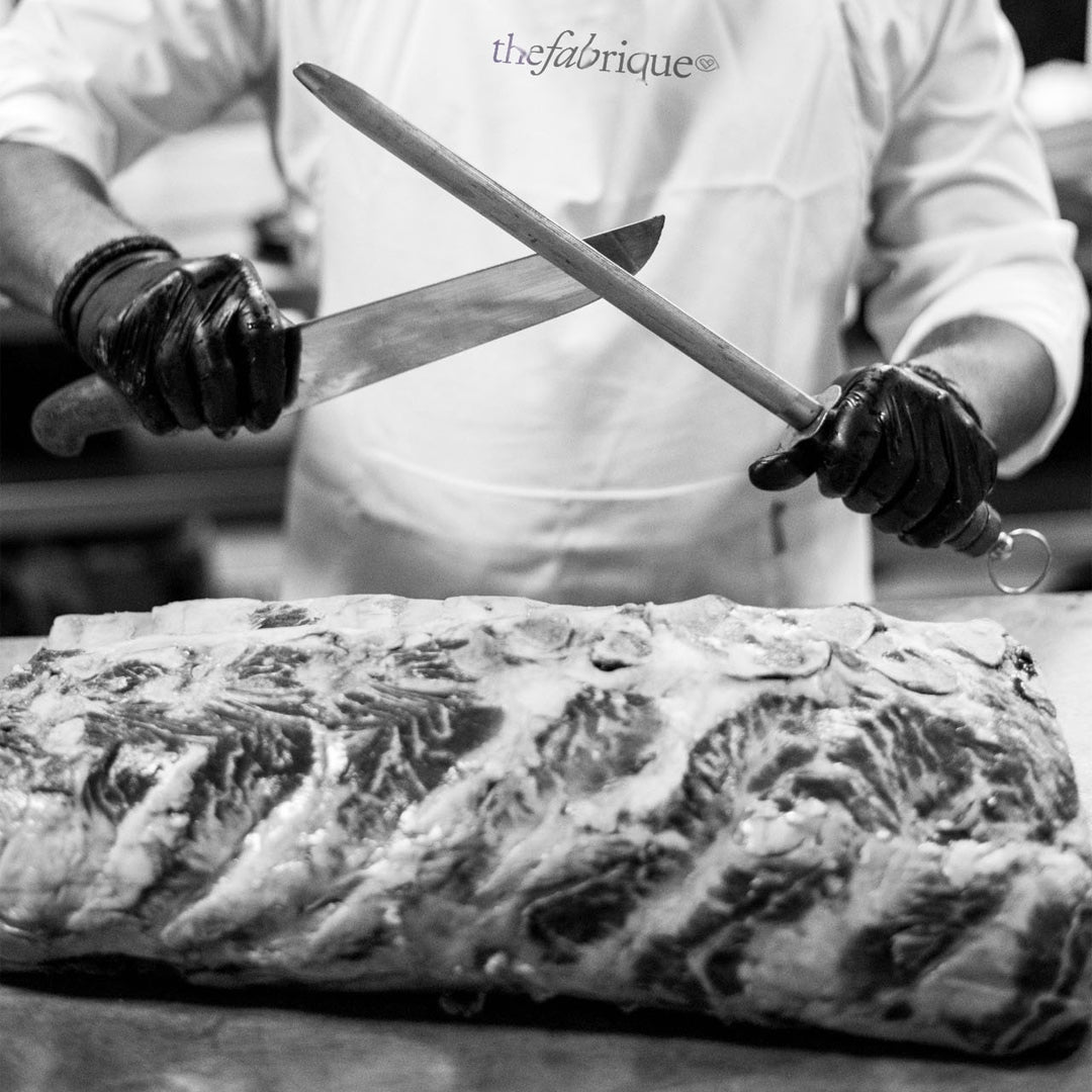 Person cutting a large piece of meat with two knives, wearing a white coat and black gloves.