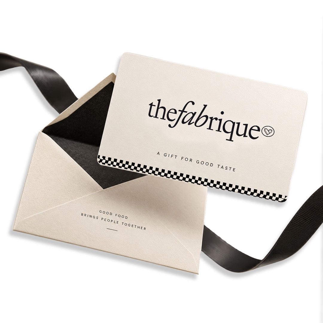 A cream-colored gift card with black text reading 'thefabrique' and 'A GIFT FOR GOOD TASTE' lies on top of a matching envelope with a black ribbon, photographed on a white background.