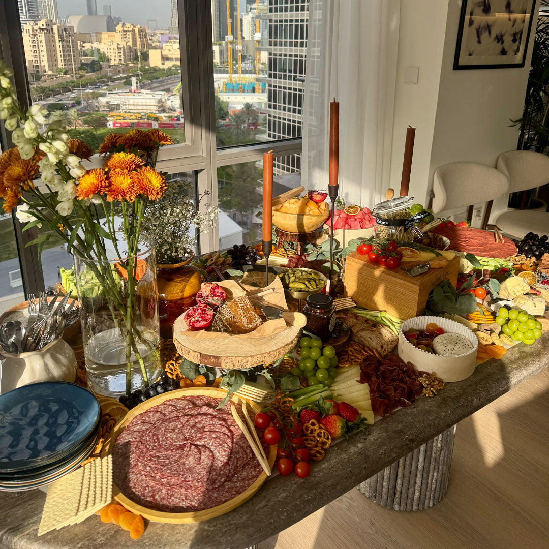 Table with a variety of food items and flowers near a window with a cityscape view.