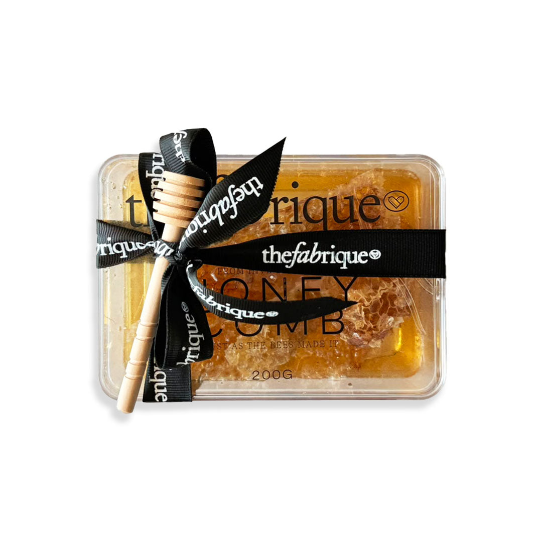 Honeycomb with a wooden dipper in a clear container, branded 'the fabrique', on a white background.