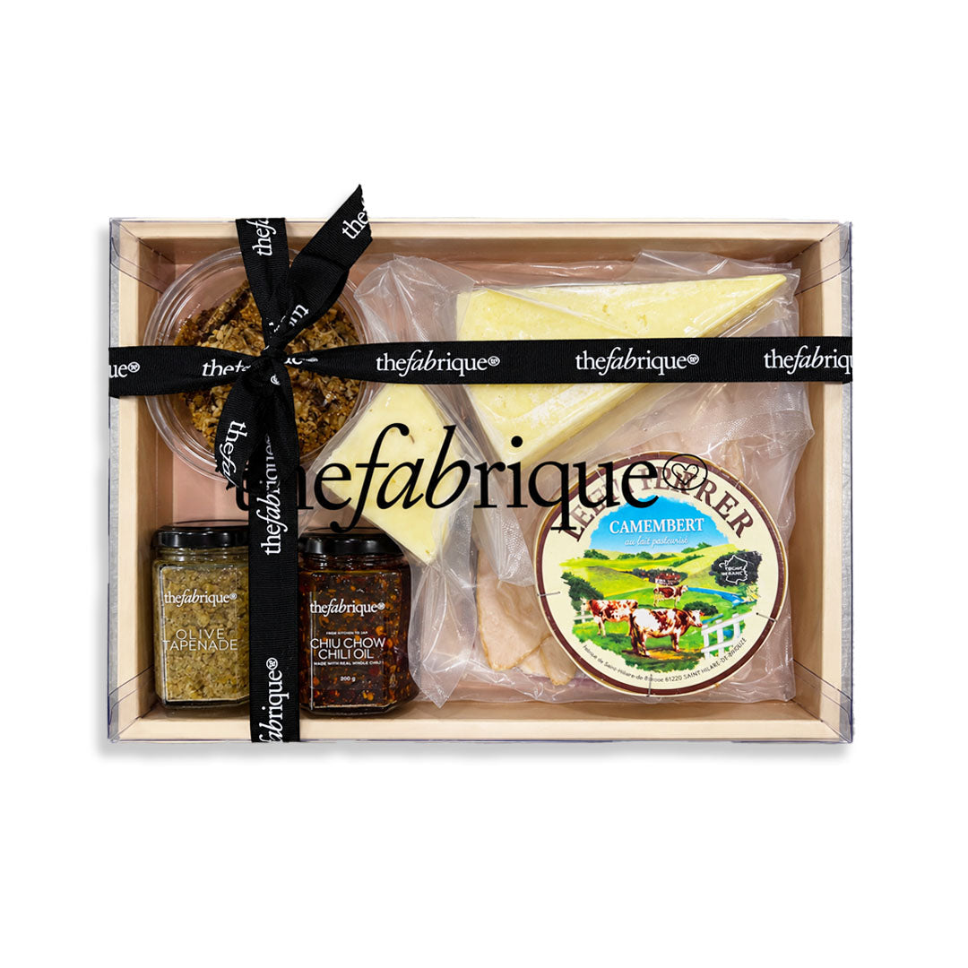 A gourmet cheese and charcuterie gift box from The Fabrique, featuring a wedge of Camembert, a small round cheese, two jars of preserves, and a black ribbon-wrapped box on a white background.