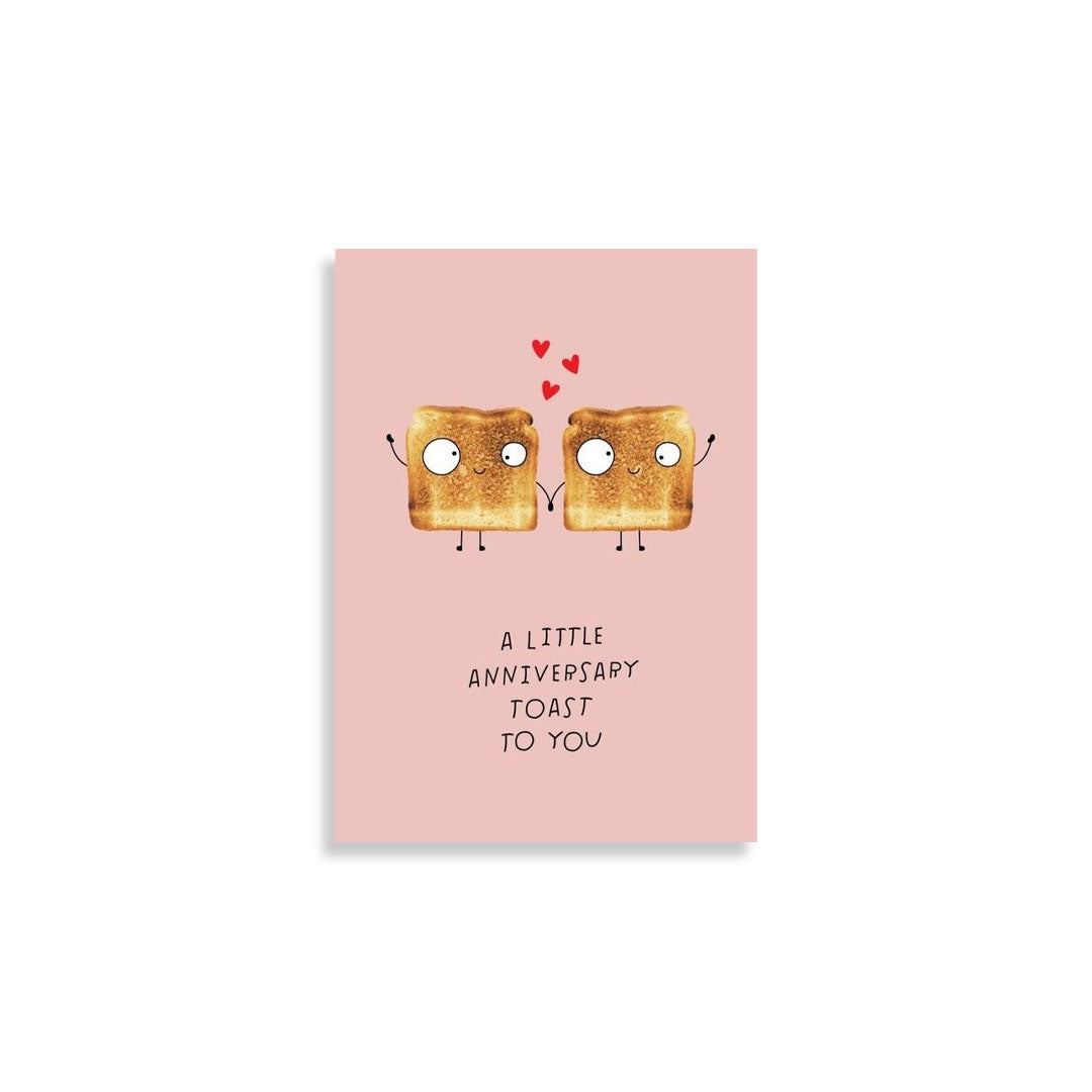 A pink greeting card featuring two cartoon slices of toast holding hands with small red hearts above and the text 'A LITTLE ANNIVERSARY TOAST TO YOU' below, shown on a white background.